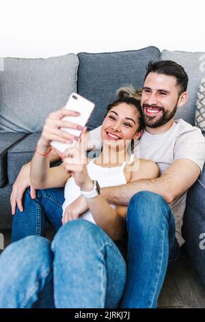 Positive ethnic couple hugging while sitting in living room at home and taking self shot on mobile phone Stock Photo