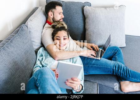 Content Latin couple of freelancers using tablet and laptop while sitting on sofa and working on remote project together from home Stock Photo