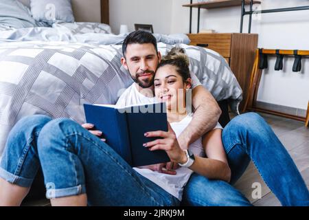 Positive ethnic couple hugging and reading book together while sitting of floor in bedroom Stock Photo