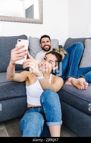 Positive ethnic couple sitting in living room at home and taking self shot on mobile phone Stock Photo