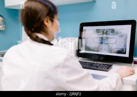 Back view of anonymous female orthodontist in mask against radiographic image of teeth on computer screen in clinic Stock Photo