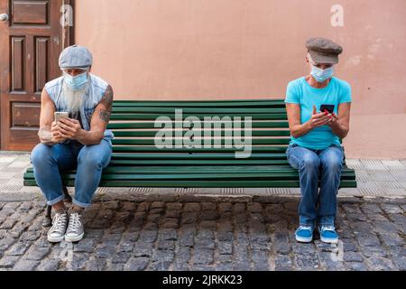 Front view of a senior couple sitting on a green bench maintaining social distance while using their smartphones Stock Photo