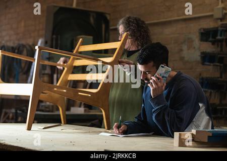Busy male joiner measuring chair detail while colleague talking on mobile phone and writing order on paper Stock Photo