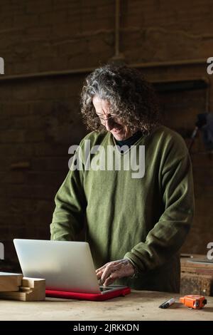 Positive mature craftsman in eyeglasses typing on laptop standing at workbench in workshop with wooden details and tools Stock Photo