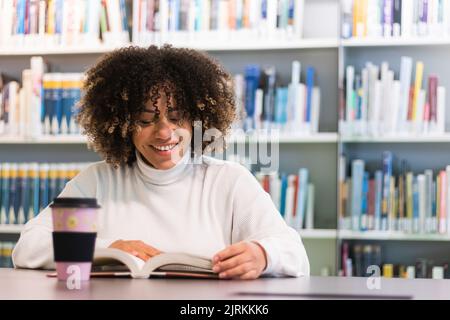 Delighted African American female student sitting at table with cup of coffee and reading book in university library while preparing for exam Stock Photo