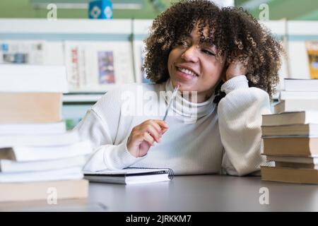 Positive African American female student sitting at table with piles of books and preparing for exam in university library while looking away in conte Stock Photo