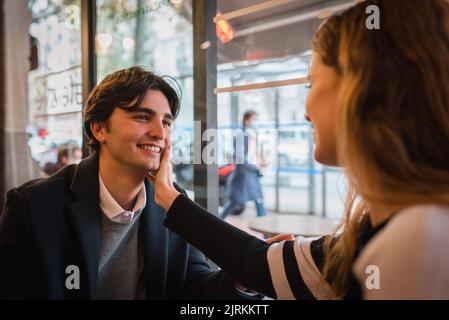 Side view of positive affectionate woman touching happy boyfriend face while looking at each other sitting at table near window in cozy cafe and enjoy Stock Photo