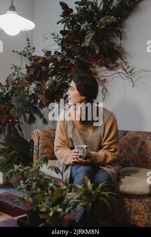 Florist resting holding a cup of coffee sitting on sofa at interior with wonderful green and brown composition of climbing houseplants Stock Photo