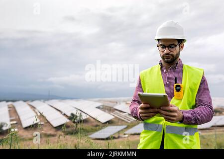 Adult man in hardhat and glasses browsing data on tablet on blurred background of solar power station and cloudy gray sky Stock Photo
