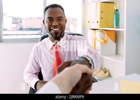 Smiling African American male boss in formal outfit shaking hand of crop anonymous coworker while looking at camera Stock Photo