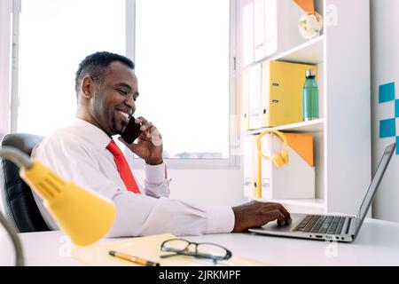 Side view of smiling black male entrepreneur talking on cellphone while surfing internet on netbook in modern office Stock Photo