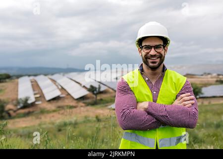 Glad male engineer with arms crossed smiling looking at camera while standing on blurred background of photovoltaic panels and cloudy sky Stock Photo