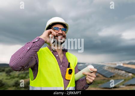 Cheerful man in hardhat holding blue prints while standing at photovoltaic panels while discussing work of solar power station on smartphone Stock Photo
