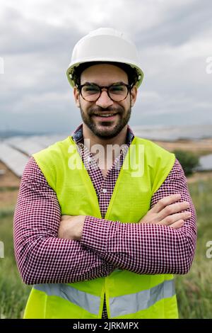 Glad male engineer with arms crossed smiling looking at camera while standing on blurred background of photovoltaic panels and cloudy sky Stock Photo
