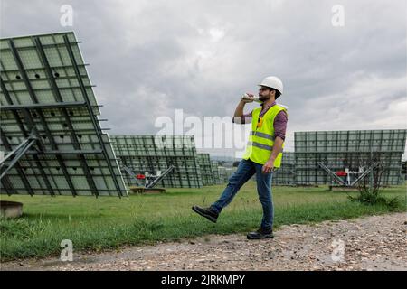 Full body man in vest and hardhat carrying draft and walking on path near photovoltaic panels on cloudy day Stock Photo