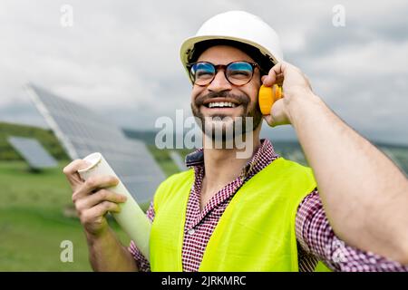 Cheerful male holding blueprint while speaking on walkie talkie against solar power station and overcast gray sky Stock Photo