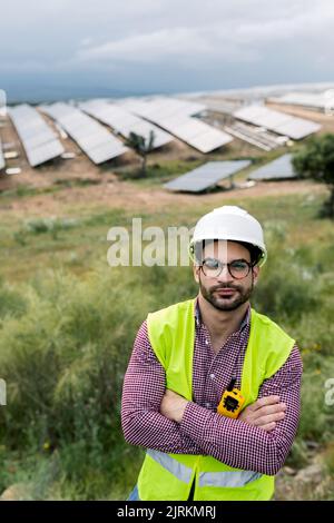 Glad male engineer with arms crossed smiling looking at camera while standing on blurred background of photovoltaic panels and cloudy sky Stock Photo