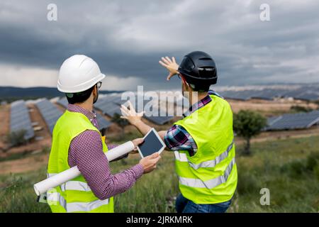 Anonymous man with tablet listening to gesticulating male colleague while discussing construction of solar power station on overcast day Stock Photo