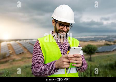 Positive man in hardhat and waistcoat smiling and browsing mobile phone while standing on blurred background of solar power station in cloudy morning Stock Photo