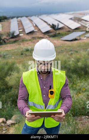 Positive adult man in hardhat and glasses browsing data on tablet on blurred background of solar power station and cloudy gray sky Stock Photo