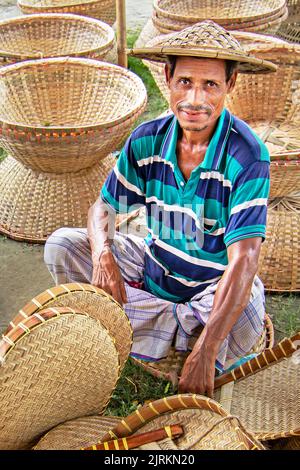 Narayanganj, Dhaka, Bangladesh. 25th Aug, 2022. A seller sits in the middle of handicrafts which are neatly arranged for sale at a weekly market in Narayanganj, Bangladesh. The baskets, Hand Fans, Fishing Traps, Caps and Handbags reflect the culture, heritage and creative expressions of people with unique artistic skills. Bangladesh earns about USD 20 million each year from the export of handicraft products. The skilled workers have honed their craft over many years. Made out of wood and bamboo, the items are characterized by the utility, sustainability and environment friendliness blended w Stock Photo