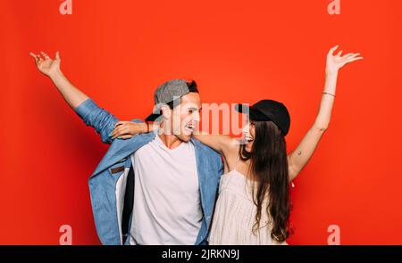 Joyful woman embracing smiling man in stylish apparel and cap while looking at each other with raised arms on red background Stock Photo