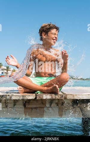 Positive teenage boy sitting on wooden quay and splashing water on sunny day in summer Stock Photo