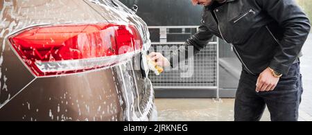 Young man washing car on carwash station outdoor. Handsome driver cleaning automobile, using sponge and soap. Stock Photo