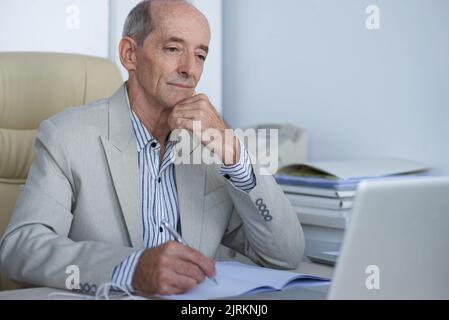 Pensive aged businesman reading something on laptop screen and taking notes Stock Photo
