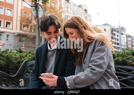 Positive young couple in love sitting on bench and discussing content on mobile phone while enjoying time together in city Stock Photo