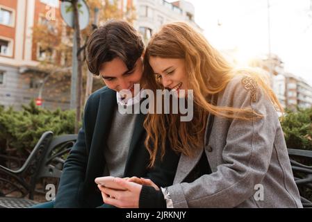 Positive young couple in love sitting on bench and discussing content on mobile phone while enjoying time together in city Stock Photo