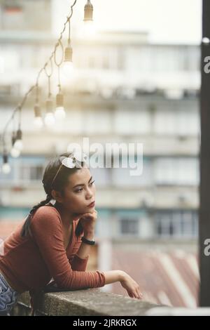 Pensive young Vietnamese woman standing on balcony and enjoying city view Stock Photo