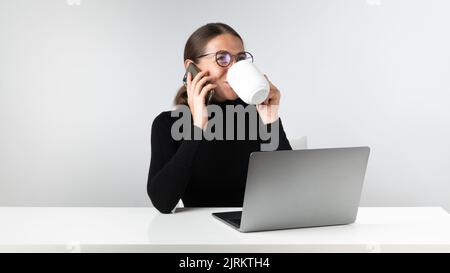 Woman at laptop talking on the phone and drinking coffee - workplace Stock Photo
