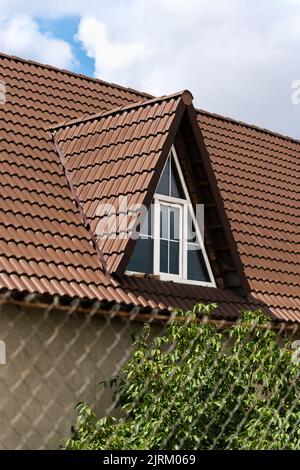 Close up shot of a triangular window on a metal tile shingles roof against blue sky with clouds. Stock Photo