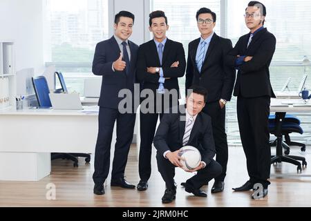 Smiling Asian managers wearing classical suits posing for photography while standing at open plan office, one of them crouching and holding soccer ball in hands Stock Photo