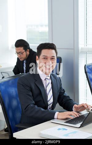 Waist-up portrait of middle-aged Asian manager wearing suit posing for photography with smile while working on promising project with modern laptop, interior of open plan office on background Stock Photo