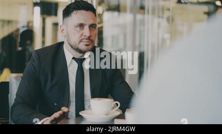 Bearded Caucasian entrepreneur gesticulating and discussing his startup plan with investor in formal wear in glassy cafe during during meeting Stock Photo