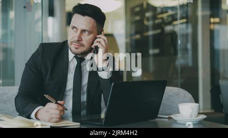 Smart Caucasian businessman sitting, talking on smartphone and writing down business information in his notepad while looking at laptop in glassy cafe Stock Photo