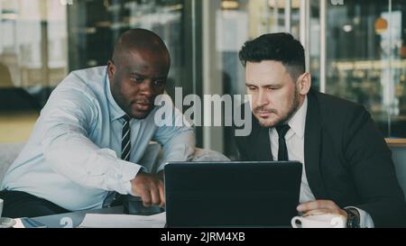 Two business colleagues looking at laptop computer and discussing their startup plan in modern office with glass walls. Bearded businessman and his partner sitting at table and talking future deals Stock Photo