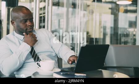 Portrait of African American businessman in formal clothes thinking, printing and working on his laptop in glassy cafe during lunch break. Coffee is on his table. Stock Photo