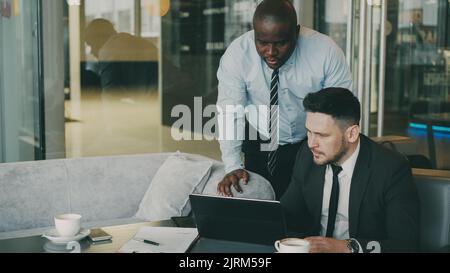 Two business partners discussing actively their startup project sitting in stylish cafe. Bearded Caucasian businessman sitting ang talking African American colleague while looking at laptop computer Stock Photo