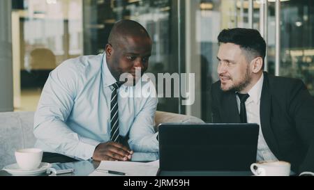 Two business colleagues looking at laptop computer and discussing their startup plan in modern office with glass walls. Bearded businessman and his partner sitting at table and talking future deals Stock Photo