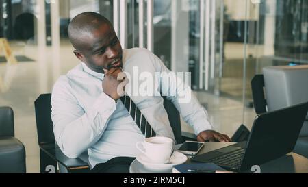 African Americanbusinessman in formal wear looking at his laptop and thinking over his business plan in glassy cafe during lunch break. He is thoughtful and thinking about project ideas. Stock Photo