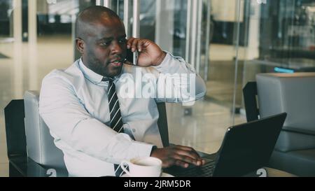 Portrait of African American businessman in formal clothes talking on his smartphone and browsing net while looking at his laptop in modern cafe during lunch time. He looks ambitious and successful. Stock Photo