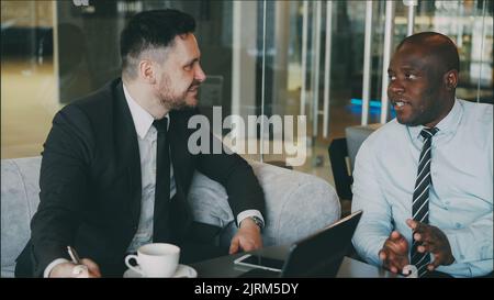 Ambitious businessmen discussing startup project in modern glassy lobby. African American entrepreneur writing in notebook and his caucasian partner in business suit using laptop computer Stock Photo