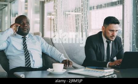 Bearded Caucasian businessman in formal clothes printing business information on his laptop and his African American partner talking on smartphone while sitting in stylish cafe during lunch. Stock Photo