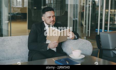 Successful Smart Caucasian businessman in formal clothes reading text book with and drinking coffee in classy cafe at lunch break Stock Photo