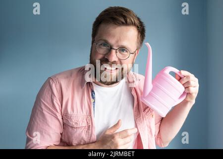 Bearded man in round glasses holds a pink baby watering can in hands and smiles while standing against a blue background. Stock Photo