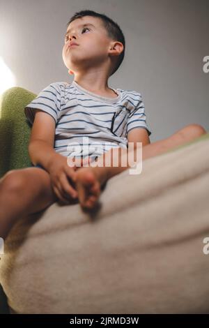 Portrait of an adorable caucasian boy sitting on the sofa at home, looking away attentively. Educational leisure pastime hobby activity. Look at TV Stock Photo