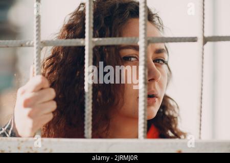 Young brunette curly woman in orange suit behind jail bars. Female in colorful overalls portrait Stock Photo